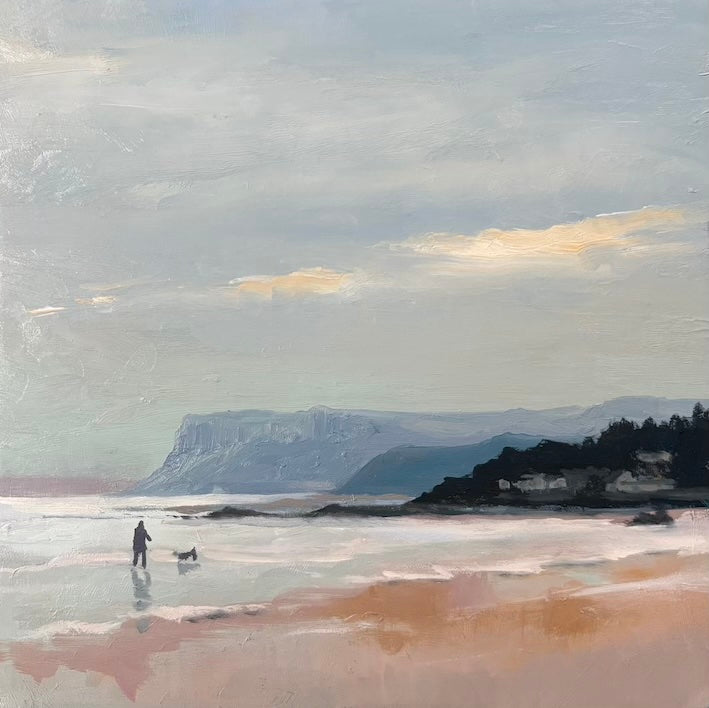 An impressionistic landscape painting of Fairhead with a pale sky, distant blue-gray cliffs, a dark shoreline with small buildings, and a pinkish beach in the foreground. A small figure stands in shallow water near the center-left walking a dog on Ballycastle strand.